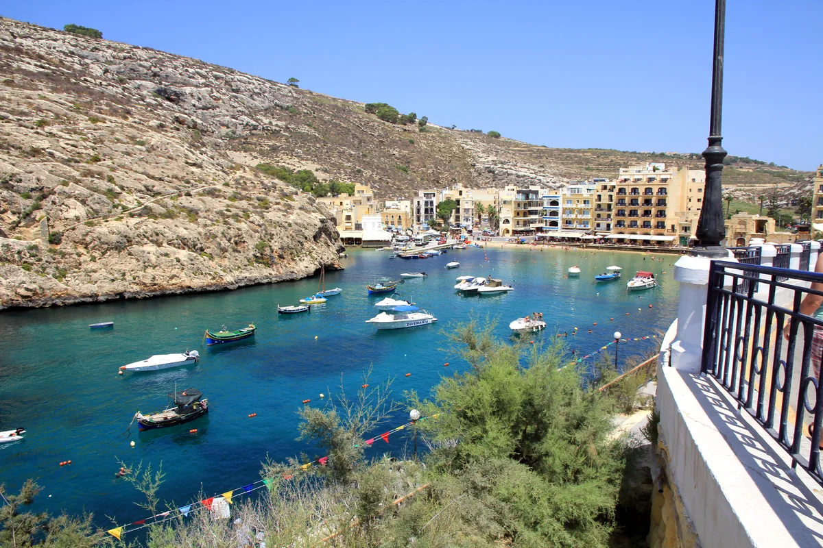 Small boats moored in turquoise Xlendi Bay beneath the cliffs and waterfront buildings.