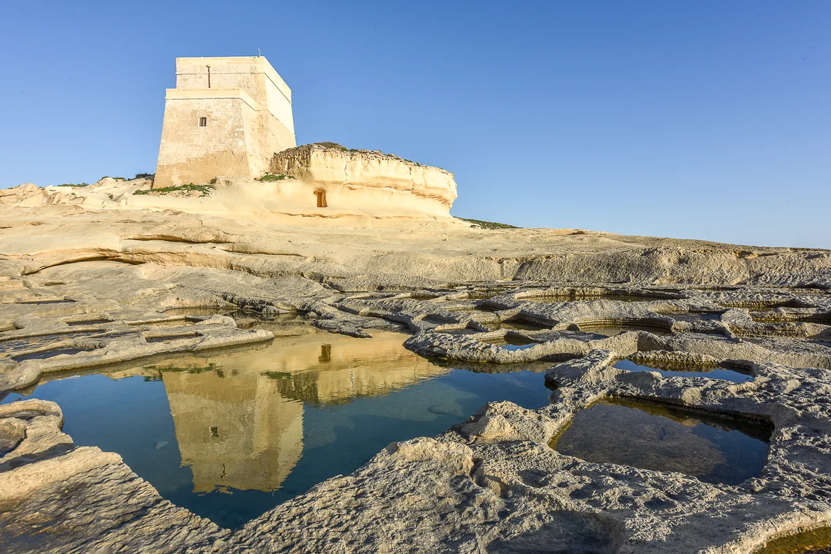 Xlendi Tower standing above limestone rock pools with its reflection in the water.